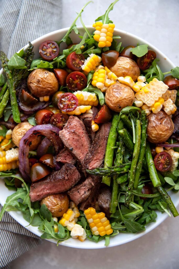 steak salad with chimichurri asparagus corn tomatoes onion in a white bowl