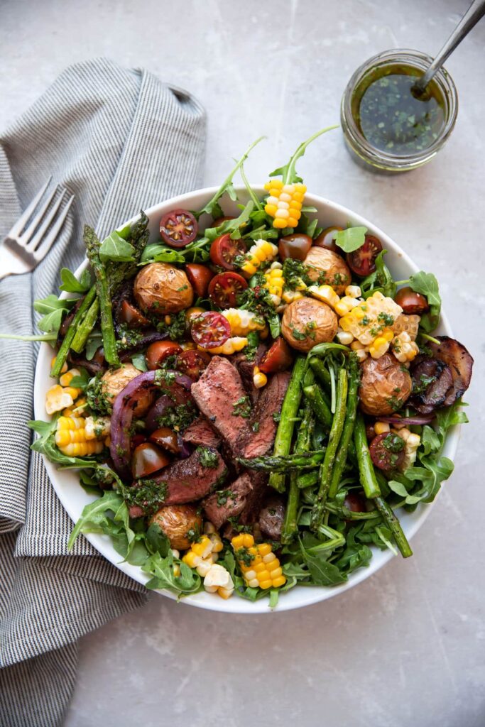 steak salad with chimichurri asparagus corn tomatoes onion in a white bowl