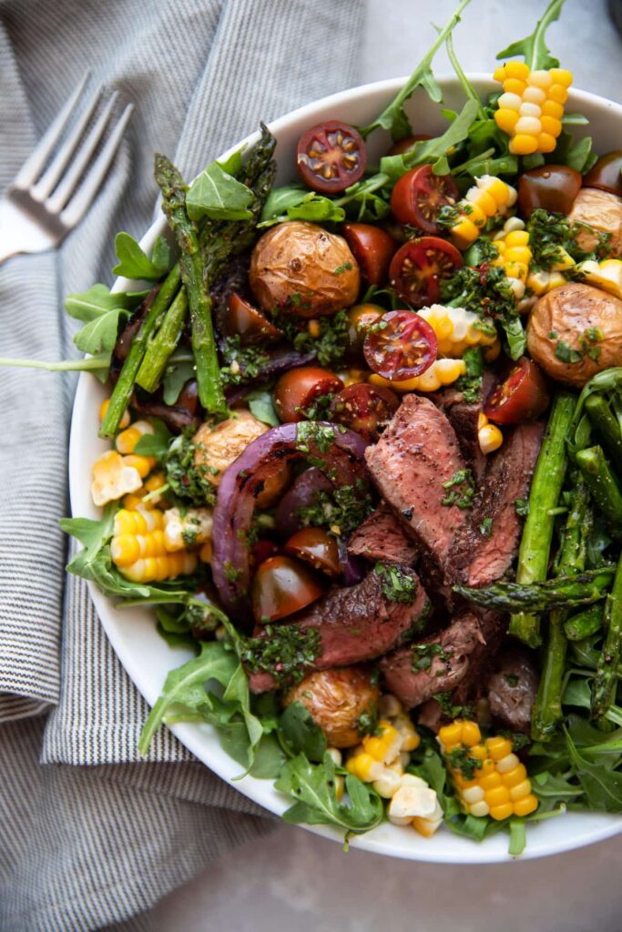 steak salad with chimichurri asparagus corn tomatoes onion in a white bowl