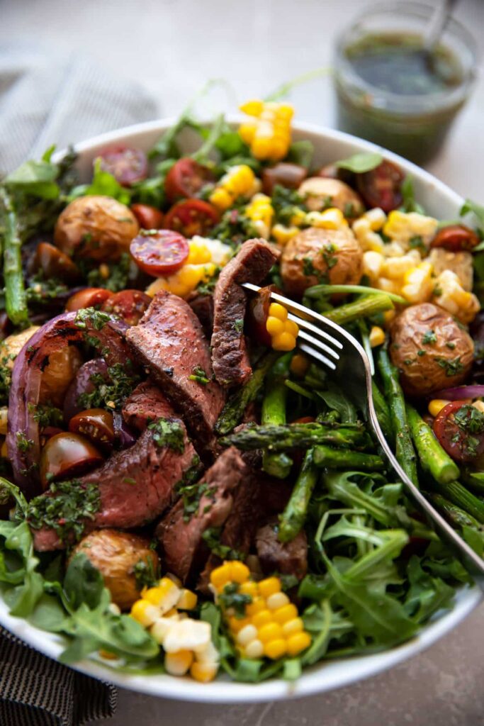 steak salad with chimichurri asparagus corn tomatoes onion in a white bowl