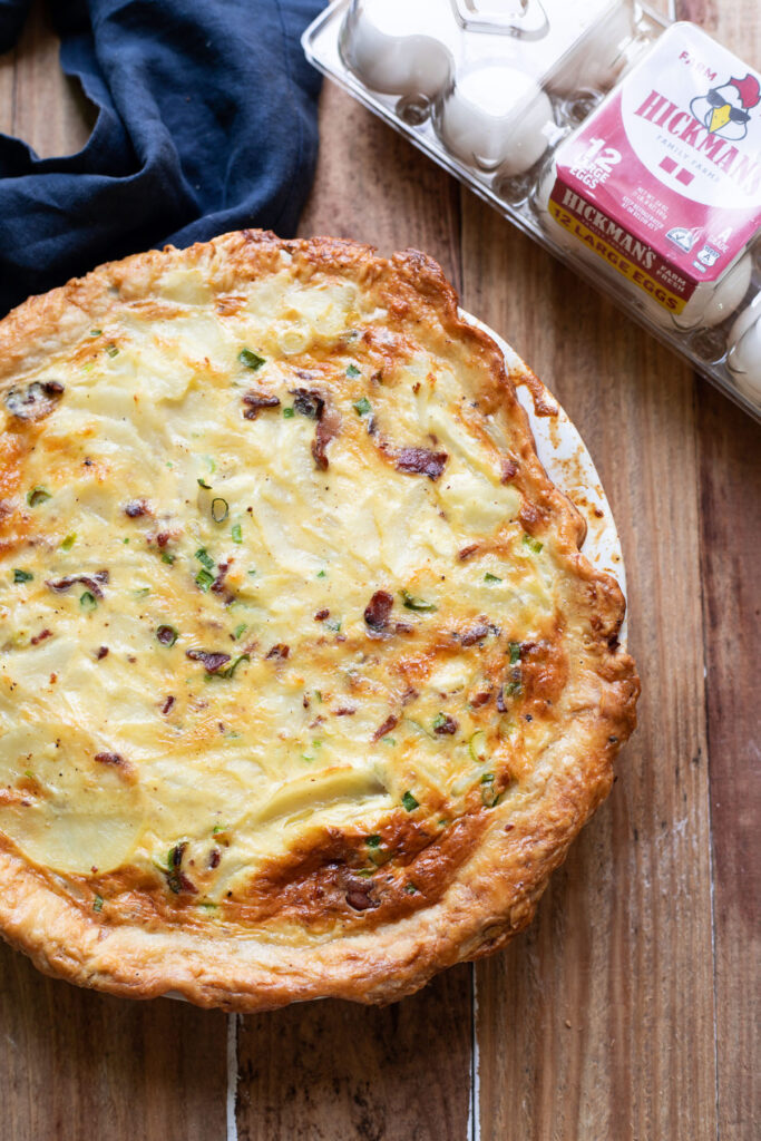 A hearty quiche made up of sliced potatoes, crispy pieces of bacon, green onion and a delicious cheesy sauce