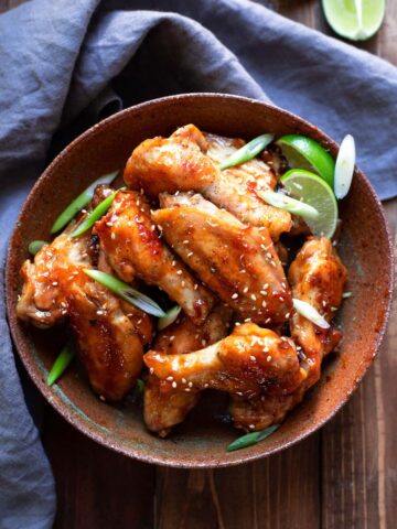 chicken wings tossed in an asian zing sauce with green onions and lime wedges in a bowl