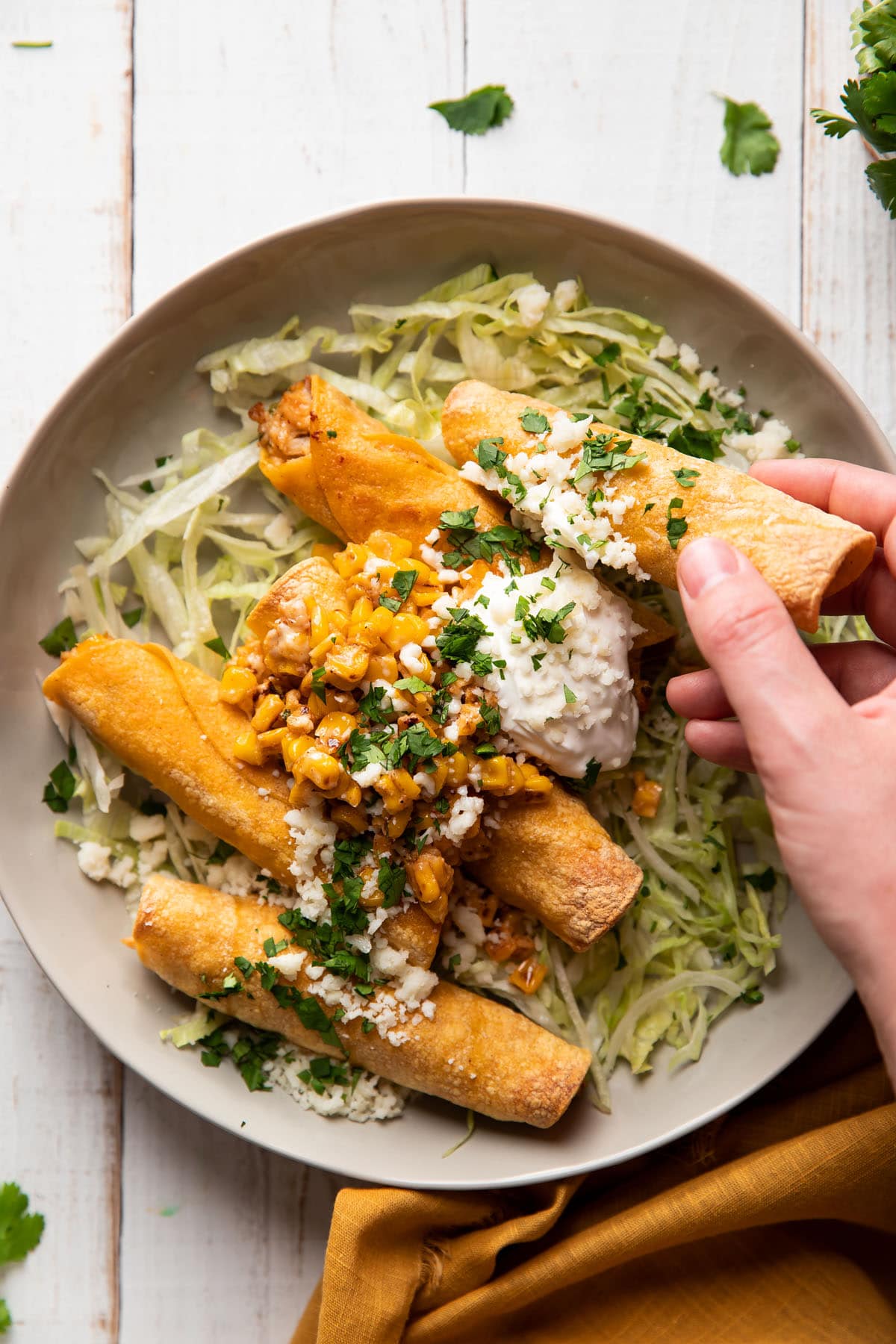 hand grabbing a taquito from a plate filled with chicken and mexican corn served with lettuce and sour cream on top