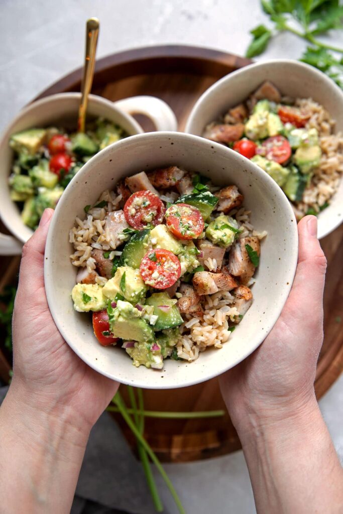 hands holding a bowl of greek marinated chicken thighs with brown rice and avocado feta dip
