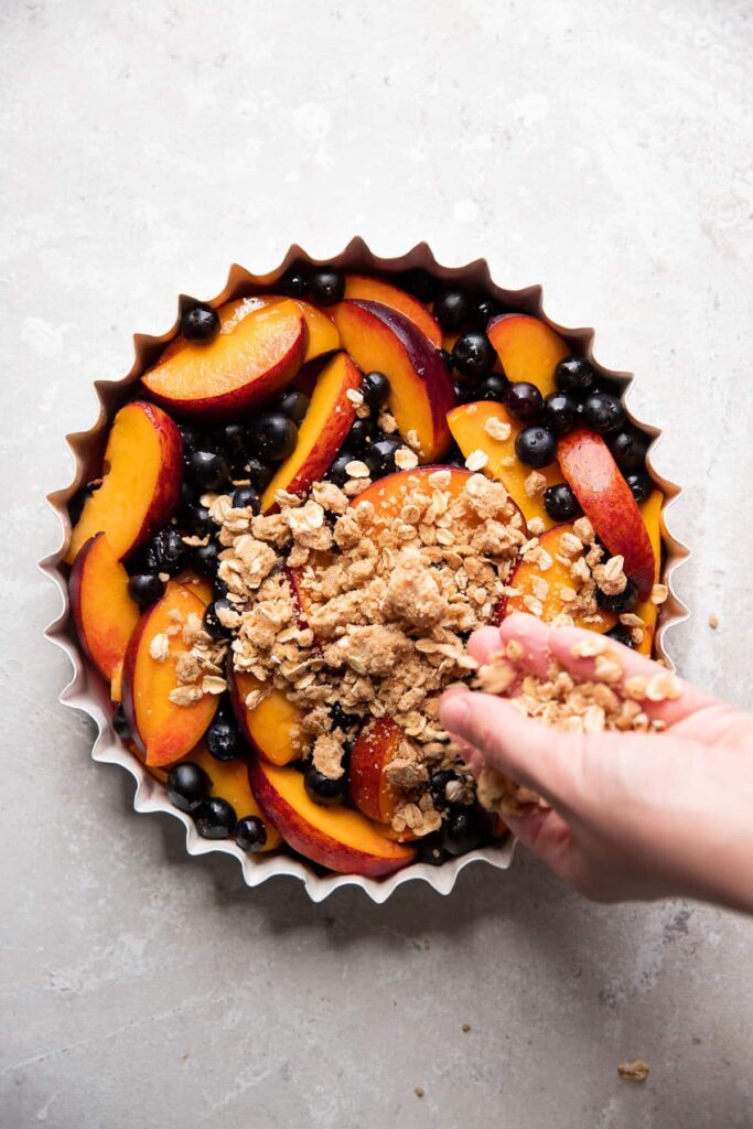 baking dish with blueberries and peaches with crisp topping being sprinkled on top