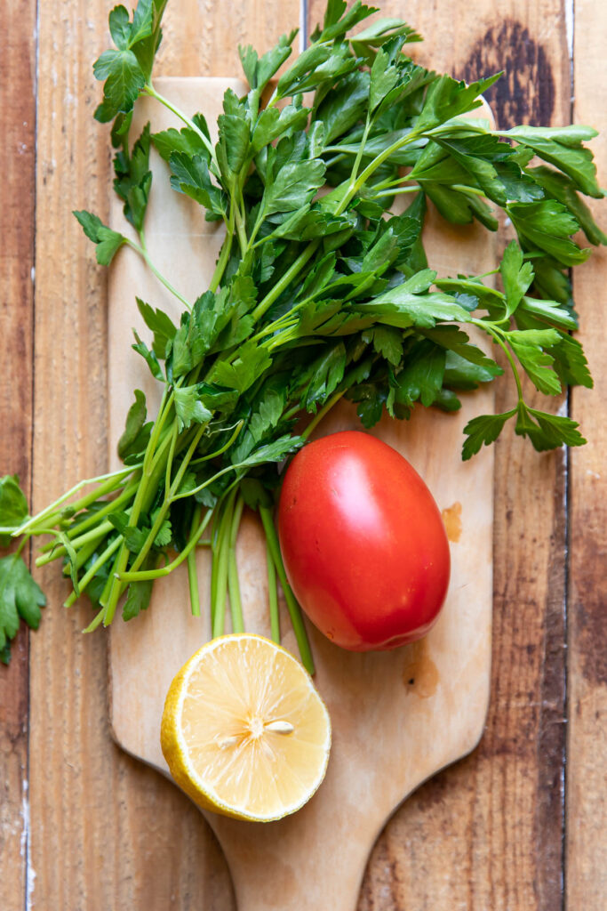 Parsley, roma tomato and half a lemon on a cutting board.