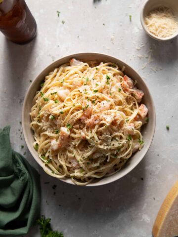 Shrimp scampi and linguine in a grey bowl, surrounded by one block of parmesan cheese and a beer.
